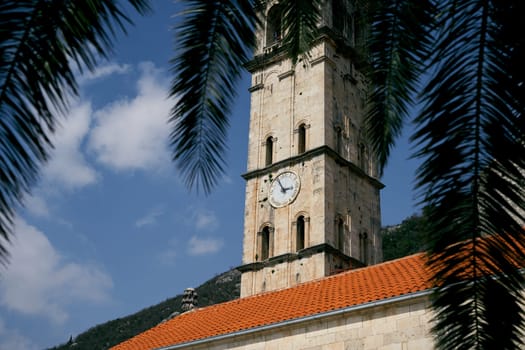 View through the palm branches to the bell tower with the clock of the Church of St. Nicholas. Perast, Montenegro. High quality photo