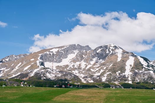 Green pasture near a small village at the foot of a mountain range covered with snow. High quality photo