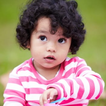 Nature, cute and girl child in a garden playing with toy on the grass on summer weekend. Adorable, playful and young kid, baby or toddler with curly hair sitting on the lawn in outdoor field or park