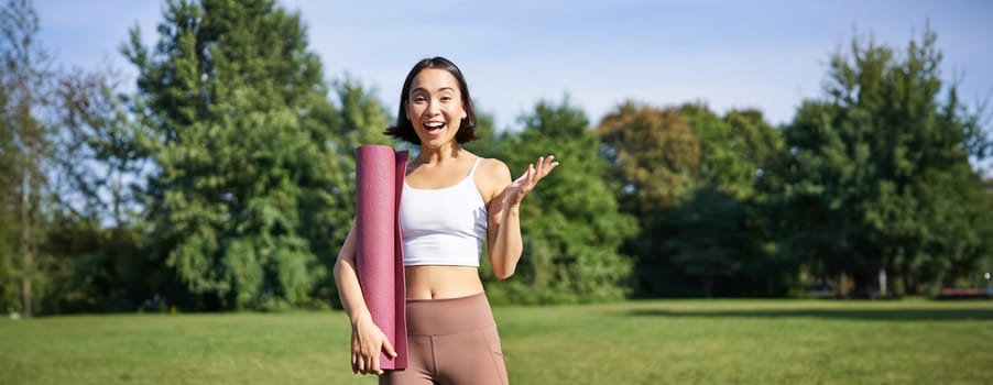 Excited asian girl in sportswear, holds rubber mat for yoga, looks surprised and happy, stands in park, workout on fresh air outdoors.