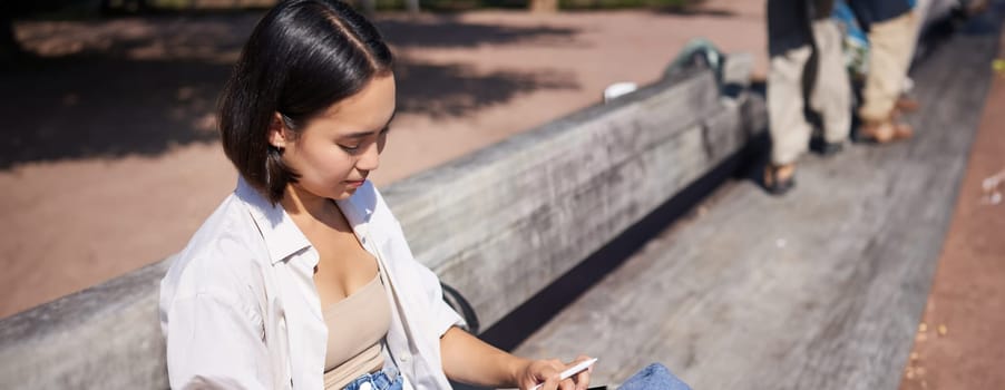 Young asian creative girl drawing with pen on graphic tablet, sitting outdoors in park, painting smth.