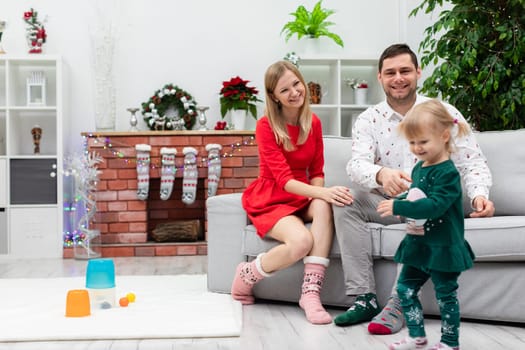 Mom, dad are sitting on a gray sofa. In front of them stands a little daughter dressed in a dark green set. A brick fireplace can be seen in the background. Christmas socks are hanging on the fireplace. The family is spending time together on Christmas.