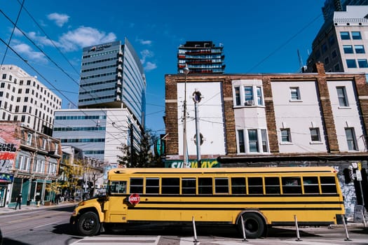 A vibrant city street featuring a yellow school bus in the foreground, contrasting older brick structures with modern high-rise buildings under a clear blue sky. High quality photo