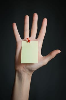 female hand with sticky office note nailed to hand with red pin. Note is blank and yellow. Dark background