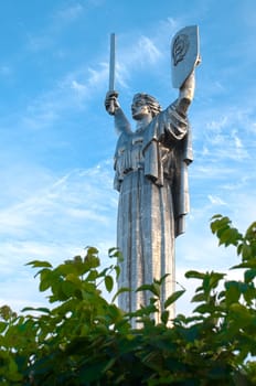 soviet motherland monument against cloudy blue sky background, green tree in foreground (Kiev, Ukraine)