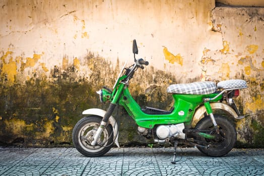 Green scooter against old house. Weathered wall as background. Urban street in Vietnam, Asia. Moped parked at moldy stonewall. Mossy surface of building. Asian lifestyle and popular transport.