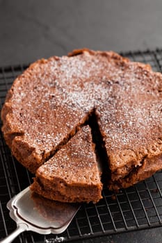 Freshly baked homemade chocolate cake on metal cooling rack with cut slice and cake server utensil on dark background
