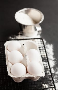 Chicken eggs in white carton on mesh tray with flour in metal measuring cup on dark background
