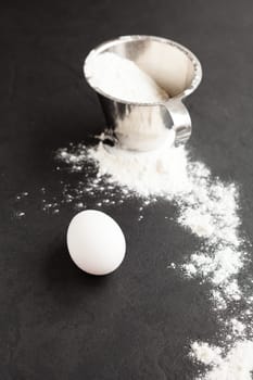 Chicken egg with flour in metal measuring cup and spread over dark background