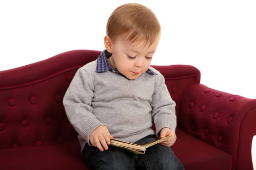 A little boy sits on a child's sofa and seems to be reading a book with interest