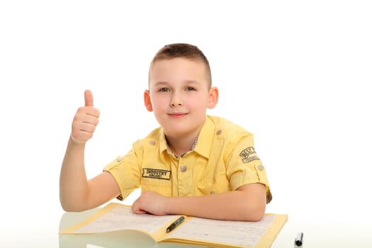 A little boy sits at a desk in front of a stapler and shows his thumb to the camera.