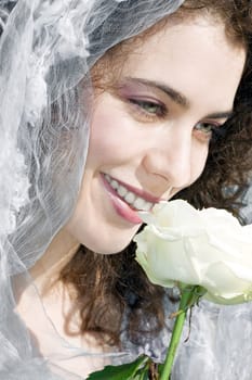 Attractive young woman with a bridal veil is smiling and smelling a white rose. Wedding and celebration concept.