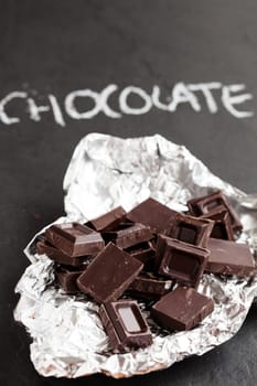 Closeup of chocolate squares in silver foil with word chocolate written in chalk on dark background