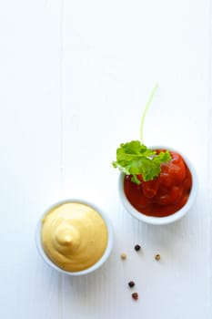 Overhead view of servings of mustard and tomato ketchup in small individual ceramic ramekins garnished with parsley on a white background with copyspace