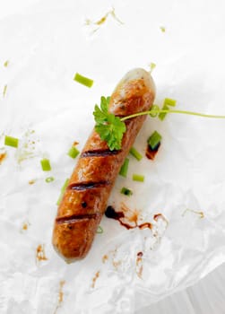 Bird's eye view of one cooked sausage on a plate with a sprig of parsley and chopped spring onions.