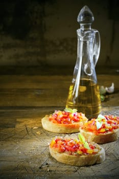 Preparing bruschetta in a country kitchen with three slices of toasted sliced baguette topped with chopped vegetables and oil with a decanter of virgin olive oil behind