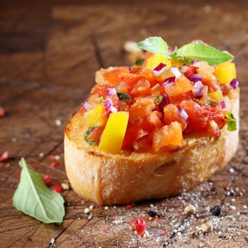 Cooking colourful bruschetta with toasted crispy sliced baguette topped with chopped tomato, onion, oil, seasoning and fresh basil on an old wooden table with scattered remnants of ingredients