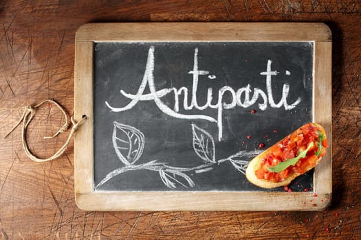 Overhead view of colourful red tomato bruschetta on an Antispasti sign handwritten on a small framed blackboard lying on a wooden background