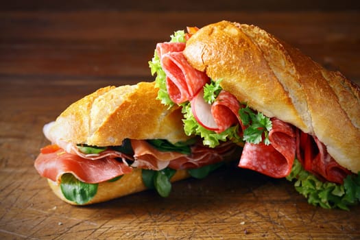 Freshly baked crisp golden baguettes filled with ham and basil or lettuce and salami on a wooden surface