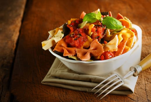 Bowl of cooked colourful Italian bow tie pasta topped with tomato sauce and basil standing on a folded napkin on an old wooden table with a fork