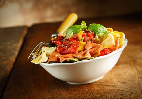 Delicious colourful Italian bow tie pasta served with tomato and basil in a white ceramic dish with a fork on an old wooden table, tilted view