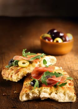 Serving of fresh traditional oven-baked Italian focaccia bread with ham, olives and rocket on a wooden table top with copyspace