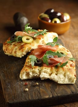 Delicous traditional oven-baked Italian focaccia bread topped with ham, rocket and olives on an old cracked wooden chopping board