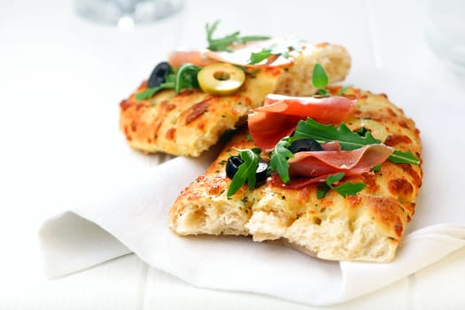 Fresh oven-baked golden focaccia bread with ham, sliced olives and fresh rocket leaves served on a white napkin