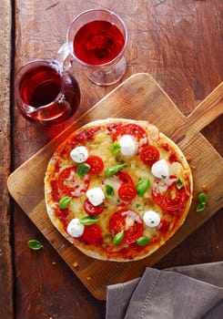 Overhead view of a colourful tricolore Italian pizza with cheese, tomatoes and fresh basil on a wooden cutting board served with a jug and glass of red wine on an old grunge wooden table. More pizza at my port.