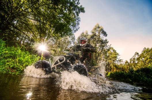 Quad rider through water stream in the forest against sunlight.