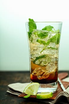 A tall glass of ice cold mujito rum cocktail with mint leaves and lime standing on a folded napkin and coaster on a wooden counter in a bar or club