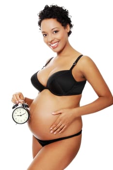 Portrait of a young beautiful pregnant woman holding an alarm clock next to her belly, isolated on a white background.