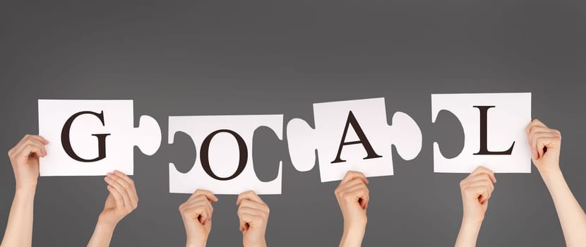 hands holding pieces of a puzzle building the word GOAL, gray background