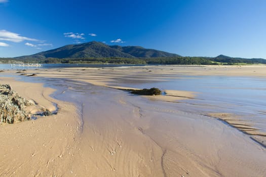 Water flowing over a sandbar toward a lake with a mountain in the background under a blue sky.