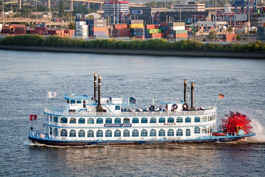 The paddle steamer Louisiana Star on the Elbe River in the Hamburg Harbor, Germany. Operated by the Louisiana Starboat Company, the boat is used for events and sightseeing primarily around the harbor area. The steamer was built in Germany to resemble the classic steamers one would associate with the Southern US, whereas the rear wheel motion is for aesthetic purposes only and the boat is driver by more conventional means. People can be seen on deck enjoying the view and Summer weather, with container terminals in the background.