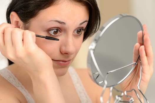 Attractive woman using mascara on her eyelashes holding handheld mirror