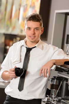 Attractive waiter leaning on espresso machine drinking cup of coffee