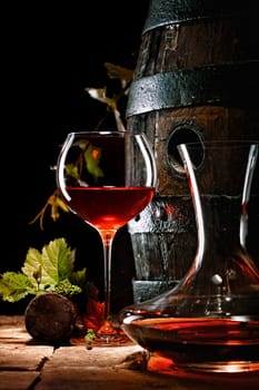 A glass of red wine next to a decanter with an oak cask in the background.