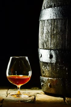 Cognac snifter with glowing golden cognac standing on old flagstones or tiles alongside an old oak barrel in a dark cellar, conceptual of aged matured cognac with copyspace