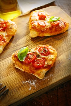 Small romantic heart shaped Italian pizzas with a freshly baked thick base topped with golden melted cheese and tomato and garnished with basil on a wooden board in the kitchen