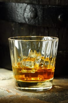 Close up of a glass of whiskey with ice and an oak barrel in the background.