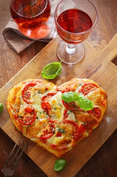 Overhead view of a tasty cheese and tomato vegetarian heart shaped pizza served on a wooden board with a glass of red wine