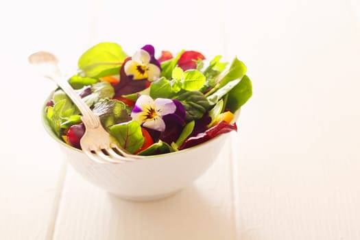 Healthy farm fresh herb salad with leafy greens and nasturtium flowers served in a white bowl with a fork on a wooden white table