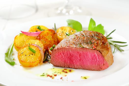 Cut through portion of delicious bloody rare to medium rare fillet steak served with crispy roasted potatoes, fresh rosemary and rocket on a white plate