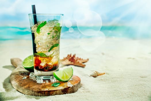 Refreshing iced rum, lime and mint mojito cocktail standing in a tall glass on an old wooden board on the golden sand of an idyllic tropical beach to celebrate a summer vacation
