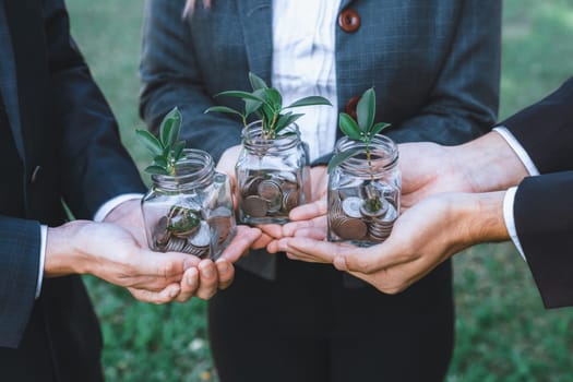 Concept of sustainable money growth investment with glass jar filled with money savings coins with businesspeople as eco-friendly financial investment nurtured with nature and healthy retirement. Gyre