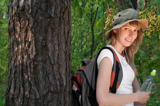 beautiful young woman standing near the tree in forest with backpack and bottle of water, smiling and looking in camera