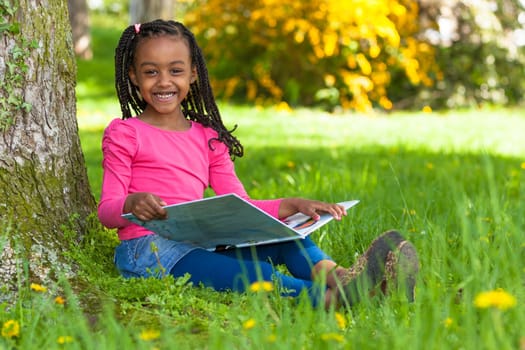 Outdoor portrait of a cute young black little  girl reading a book - African people