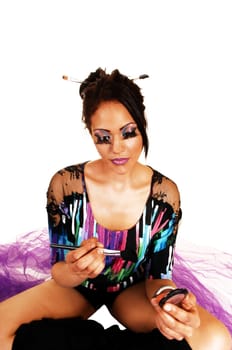 A young woman sitting on lilac twill on the floor with crossed legs and black long boots putting on her makeup, for white background.