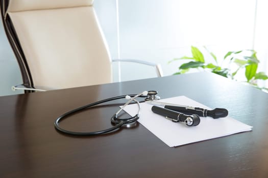 Doctor office table desk and beige chair with stethoscope otoscope and white paper